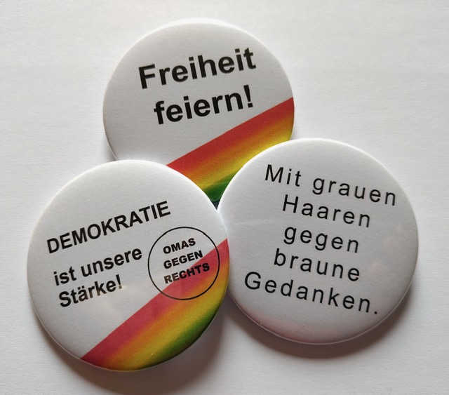Drei Buttons mit den Sprüchen ‚Freiheit feiern!‘, ‚Demokratie ist unsere Stärke!‘ und ‚Mit grauen Haaren gegen braune Gedanken.‘, teils mit Regenbogenstreifen.
