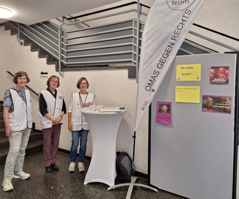 Informationsstand der Omas gegen Rechts mit Roll-up-Banner und Infomaterial, zwei Personen am Tisch.