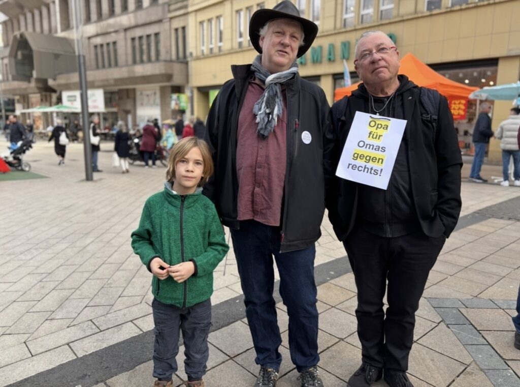 Zwei Männer und ein Junge in der Dortmunder Innenstadt. Einer der Männer trägt ein Schild mit der Aufschrift ‚Opa für Omas gegen Rechts!‘